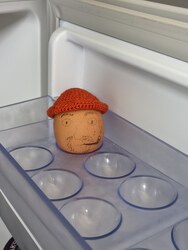 An egg wearing a small orange crochet beanie, sitting in the fridge egg tray with a face drawn on it