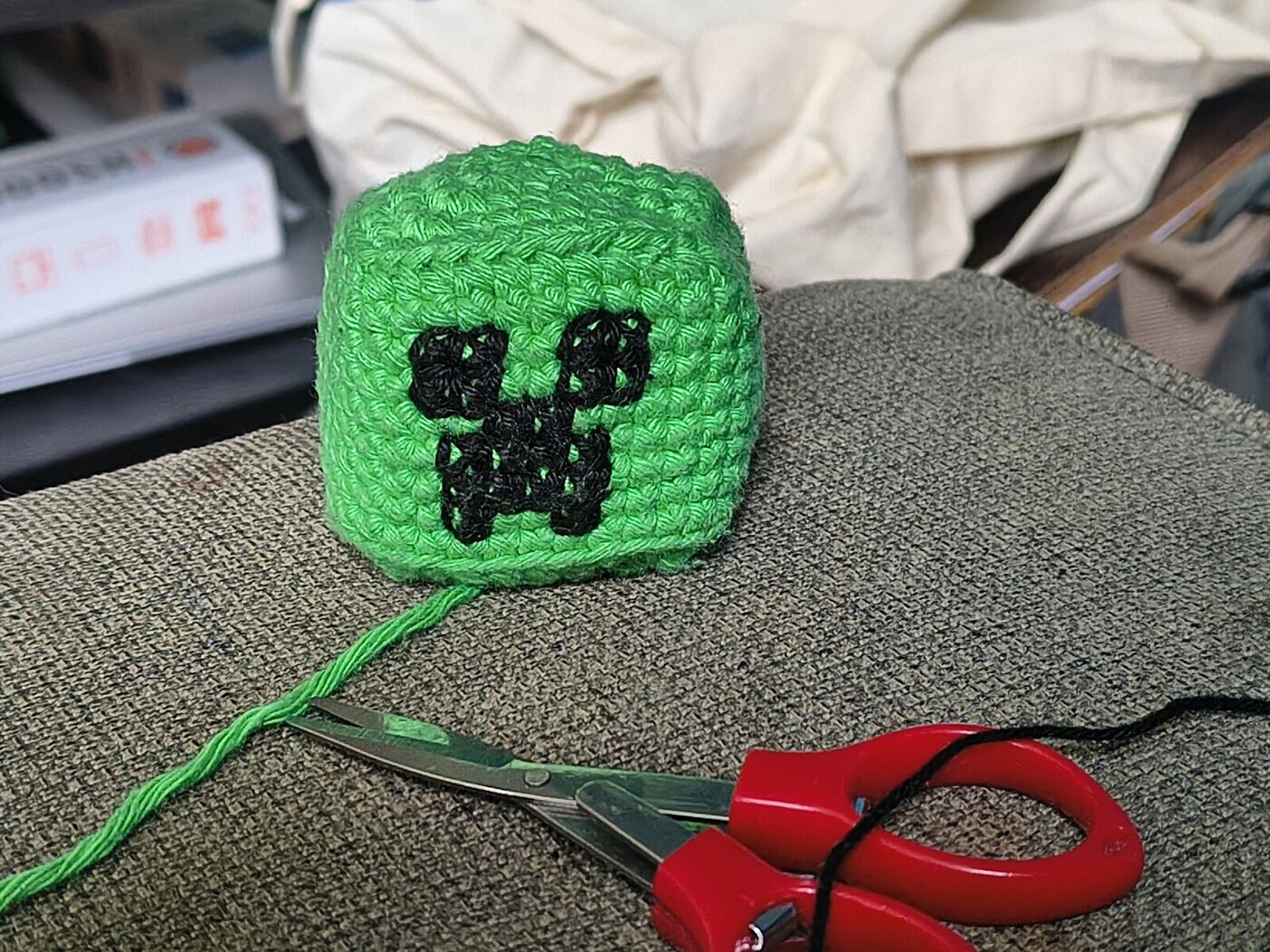 Work in progress: the green crochet cube head with the Creeper face embroidered in black yarn, next to red scissors, the green yarn tail and a needle threaded with black yarn