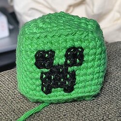 Close-up of a green crochet cube with the black Creeper face embroidered on the front